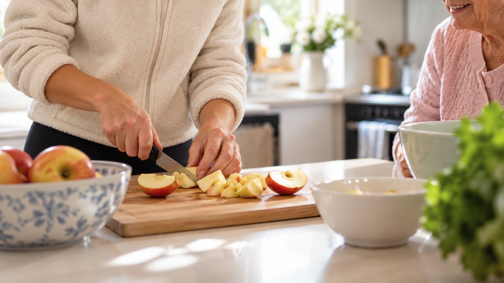 A helper slicing apples on a wooden board in a bright kitchen while an older lady smiles beside her
